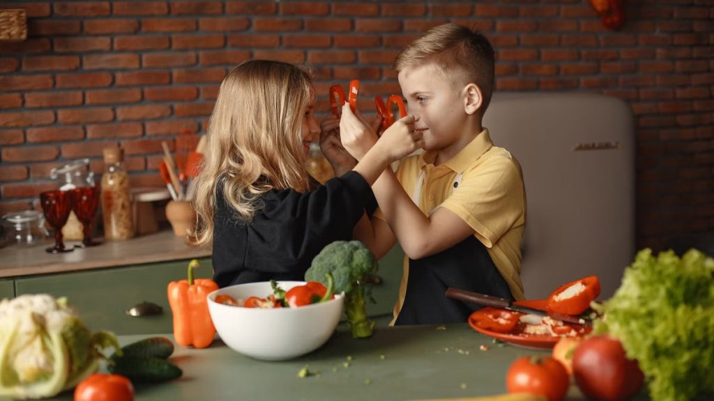 Playful boy and girl wearing apron preparing healthy vegetarian salad together at table with ingredients and bowls while looking at each other through slices of pepper
