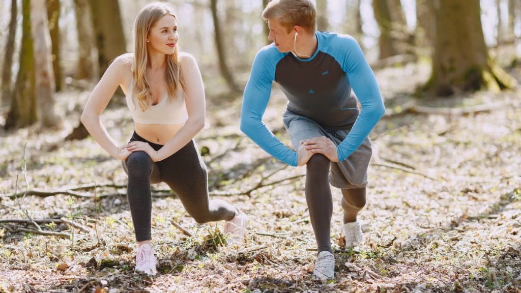 A couple engages in outdoor stretching exercises in a sunlit forest setting, promoting wellness and fitness.